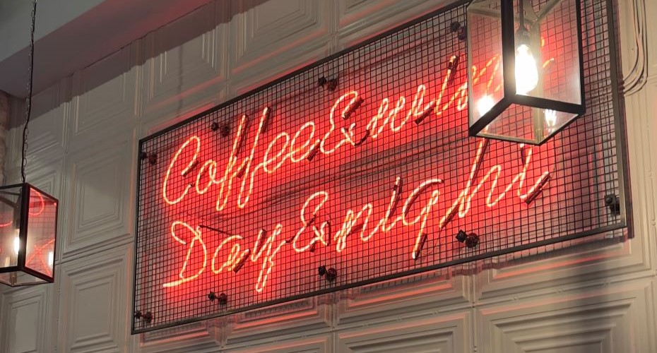Red neon sign surrounded by a black wire cage on a white panelled wall that says 'Coffee & Wine, Day & Night'