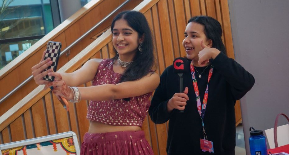Two female students posing for a selfie next to a wood panelled set of stairs. One student is wearing an embellished red top and skirt and the other is wearing a black top and Students' Guild lanyard.