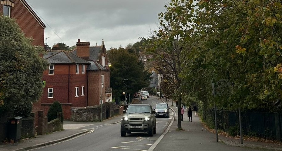A few cars driving on a residential street on Exeter, which is surrounded by brick houses, a few trees and two pedestrians, all under a cloudy sky