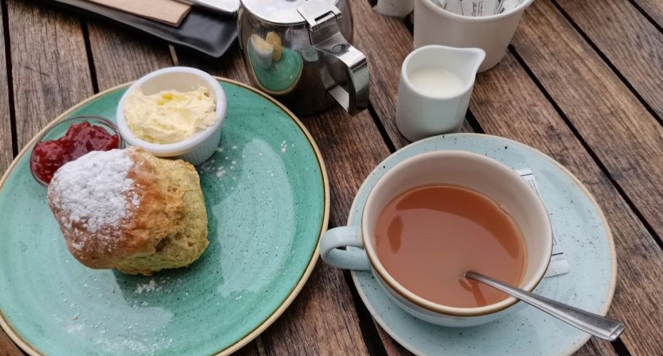 Cup of tea, milk, sugar, a metal teapot and a scone on an outside table