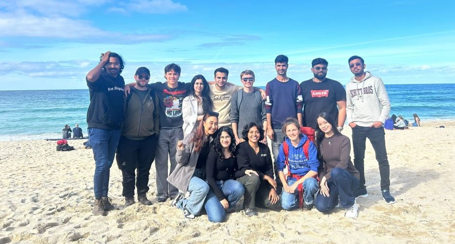 A large group of students posing and smiling for a photo on a white sand, sunny beach