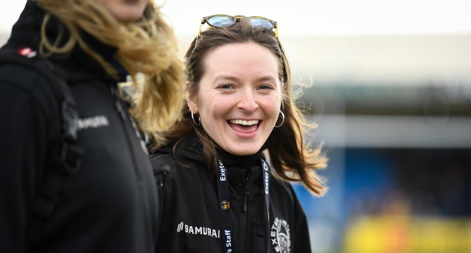 Photo of a female member of Exeter Chiefs staff smiling at the camera wearing a branded jacket and lanyard