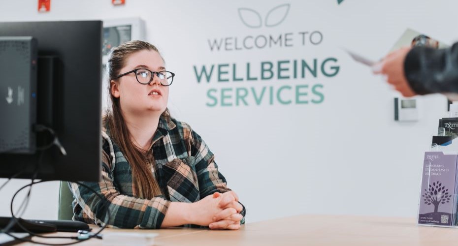 Woman sat at a reception desk in front of a green 'Wellbeing Services' sign