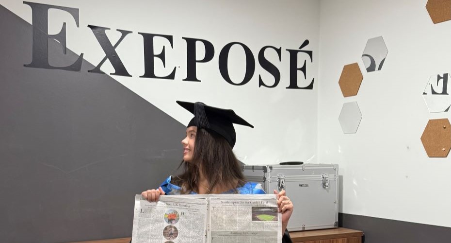 Female student wearing a graduation cap and holding a newspaper under a sign that says 'Exposé'