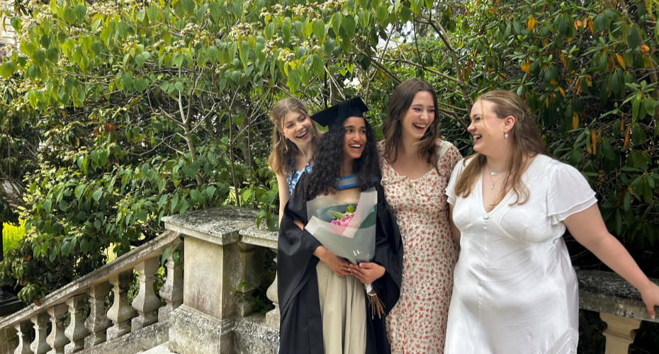 A group of female students, including one in a graduation robe, smiling together on a outdoor stone staircase